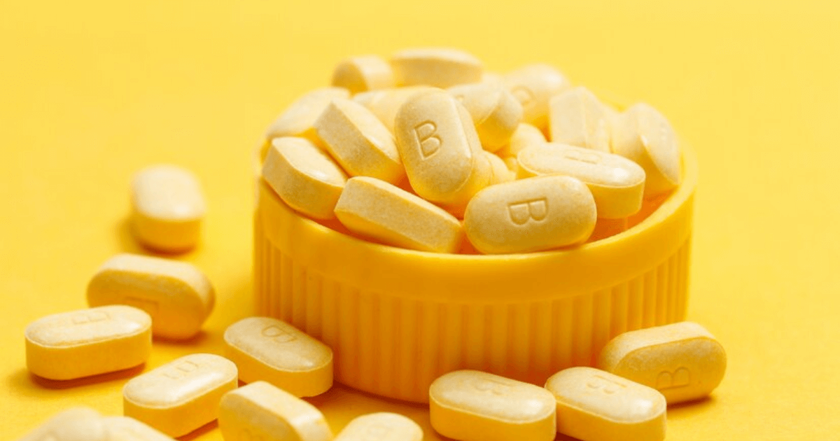 A bottle of Vitamin B Complex capsules surrounded by fresh spinach leaves, almonds, eggs, and avocado on a white marble countertop, symbolizing natural sources of B vitamins.